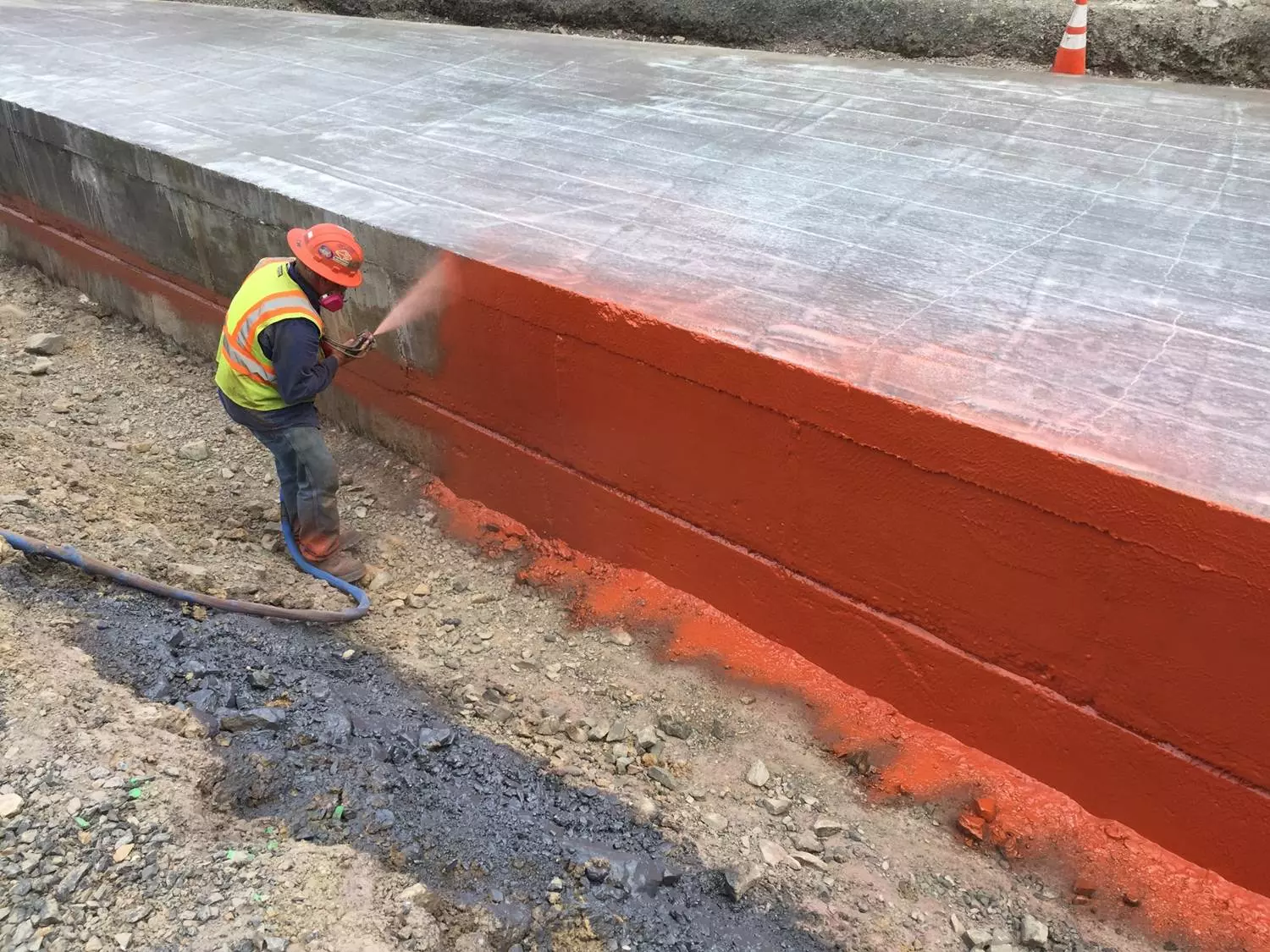 An image of one construction worker using a sprayer to apply an orange coating to a bridge.